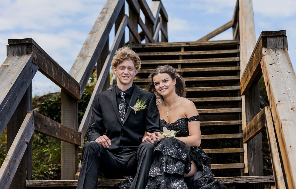 Prom couple in matching black formal wear posing on wooden staircase outdoors.