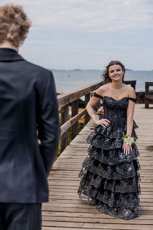 Smiling teen in black tiered sequin prom gown posing on wooden pier with ocean backdrop.