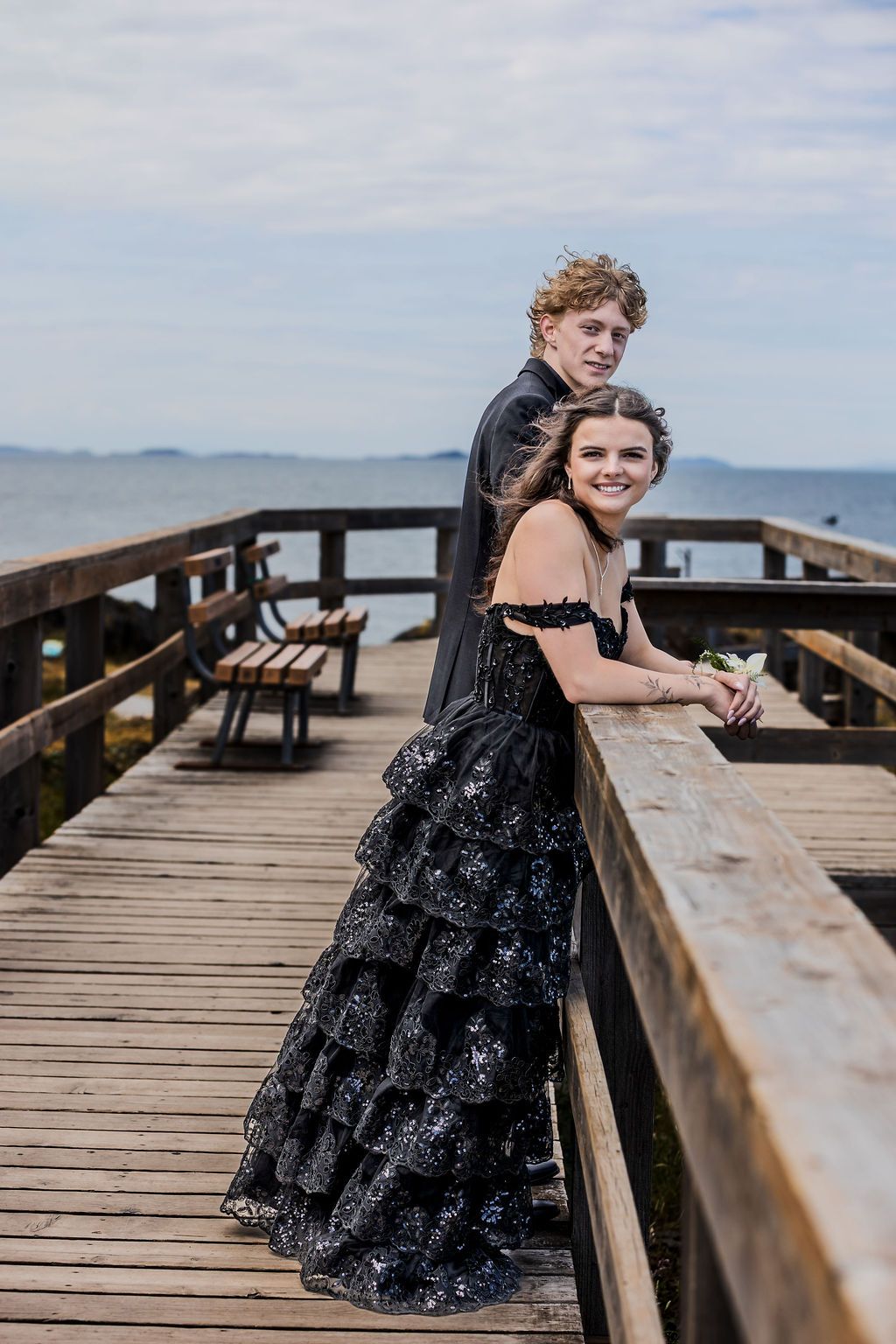 Prom couple in black formal wear posing on a wooden waterfront pier with ocean views.