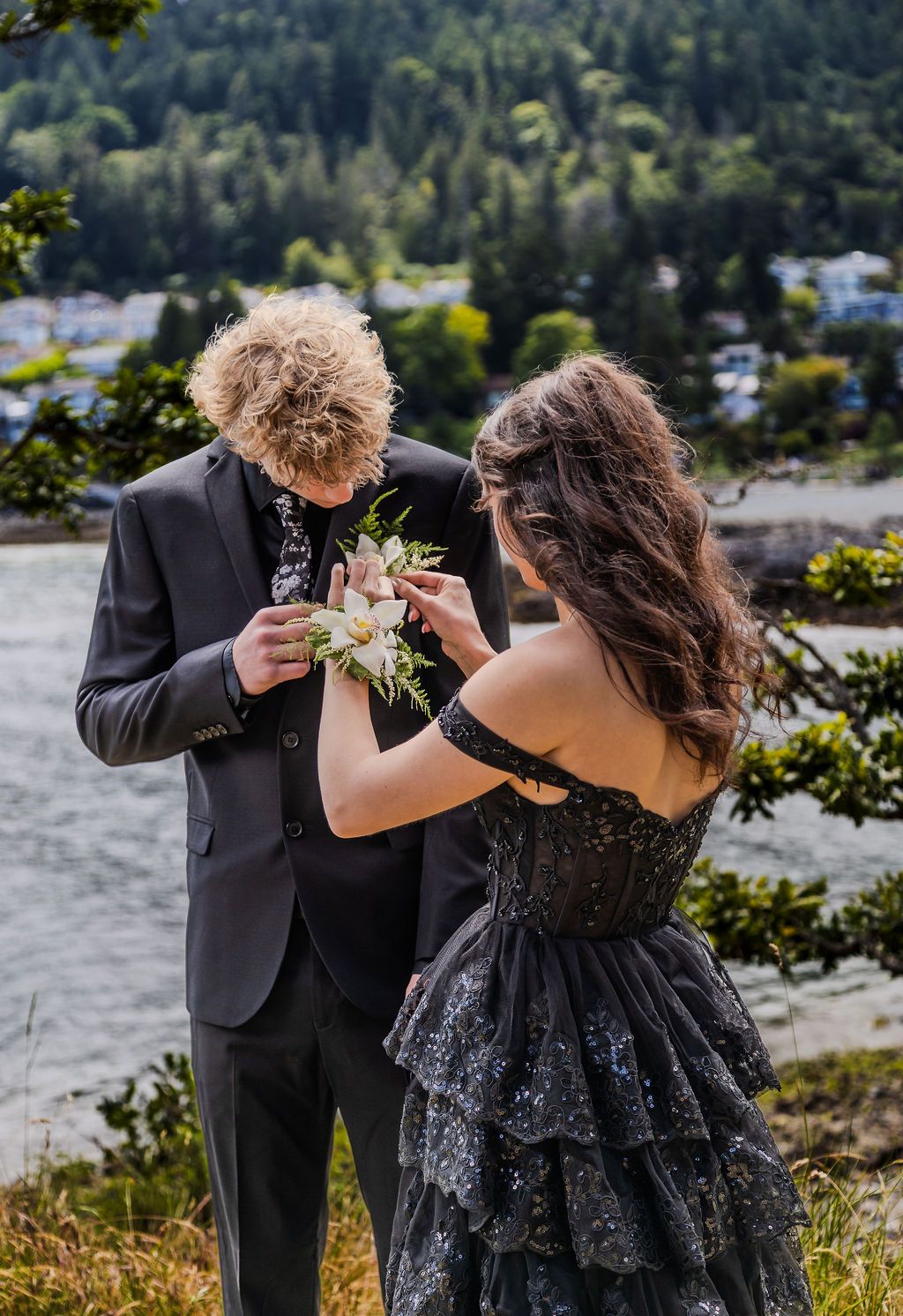 Young woman in black sparkle gown pins a boutonniere on her date's suit lapel outdoors.