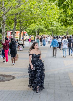 Teen in black tiered sequin off-shoulder prom gown walking through a busy tree-lined plaza.