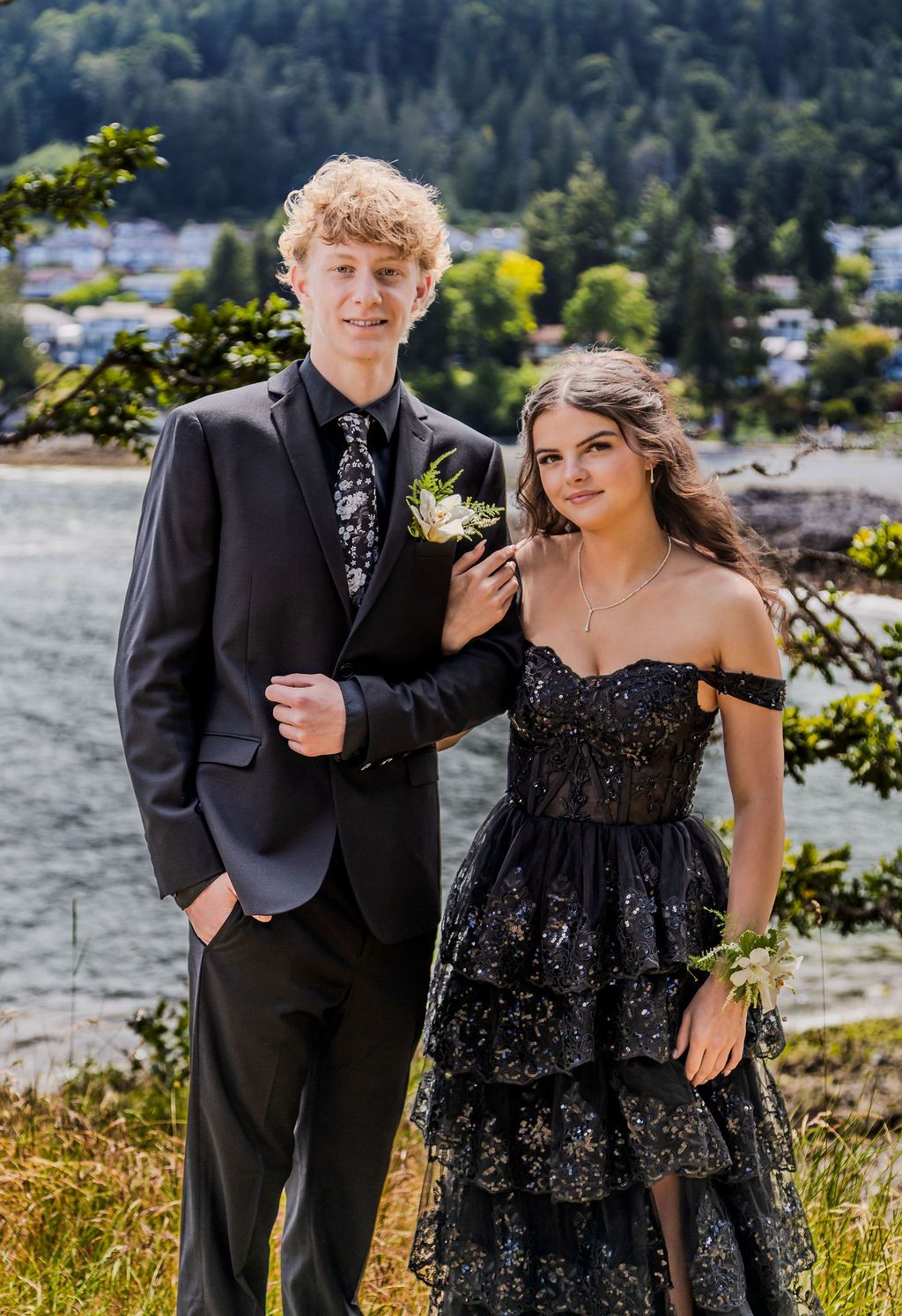 Young couple in black prom attire pose outdoors with a scenic waterfront and forest backdrop.