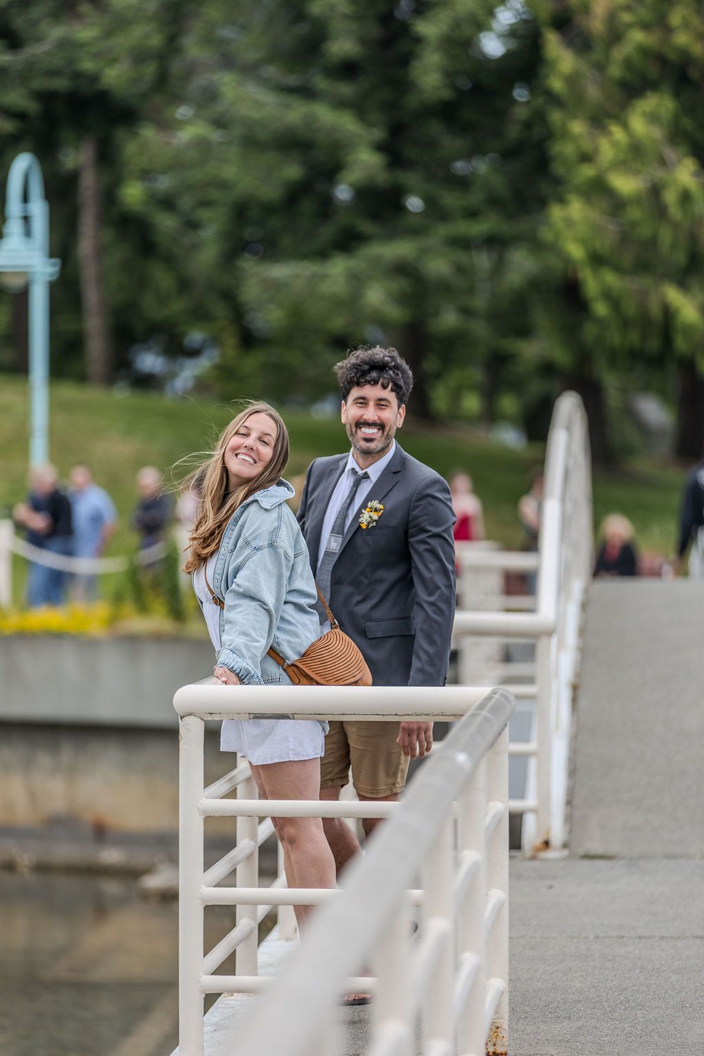Smiling prom couple leaning on white railing of a waterfront bridge surrounded by greenery.