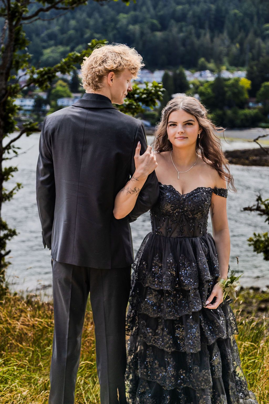 Young prom couple in black formal attire share a moment outdoors by a scenic waterfront.