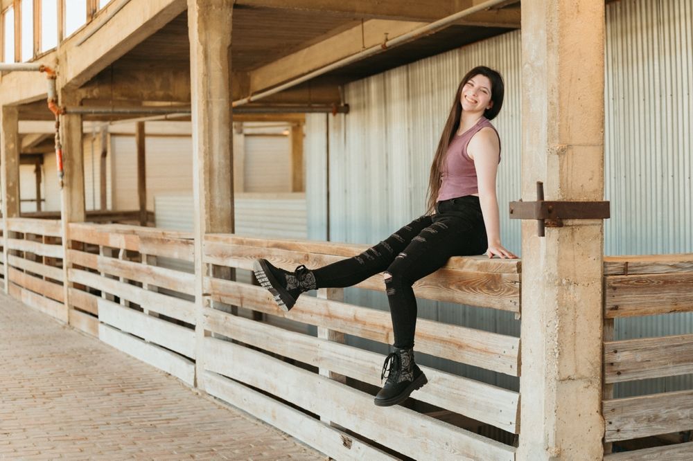 Young woman in black leggings and boots posing with leg raised on wooden fence at rustic outdoor structure.