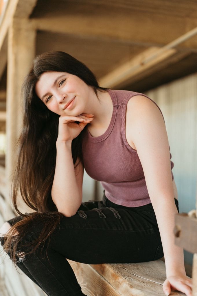 Young woman with long dark hair poses on wooden bleachers in a mauve tank top and black ripped jeans.