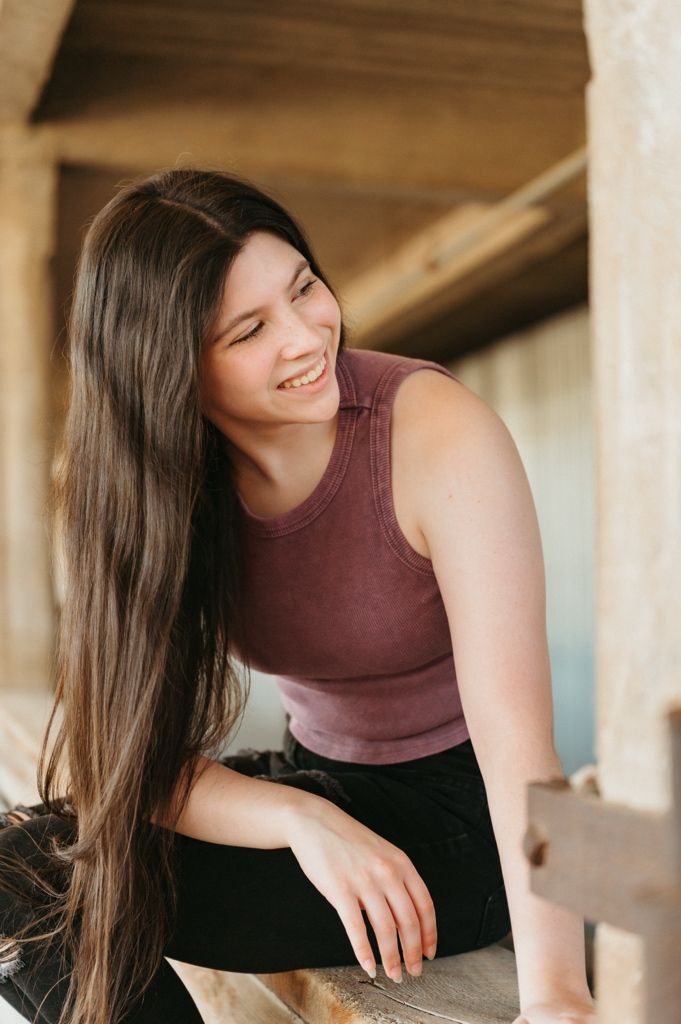 Smiling young woman with long dark hair wearing a mauve tank top sitting on a wooden fence outdoors.