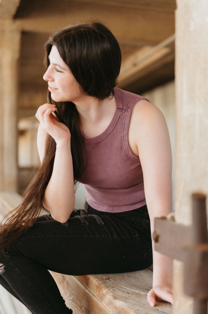 Young woman with long dark hair sits thoughtfully on a wooden beam wearing a mauve tank top and black jeans.