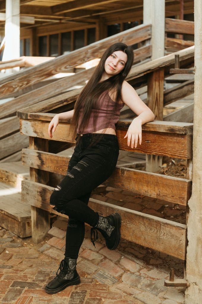 Young woman in black ripped jeans and boots poses on rustic wooden bleachers outdoors.