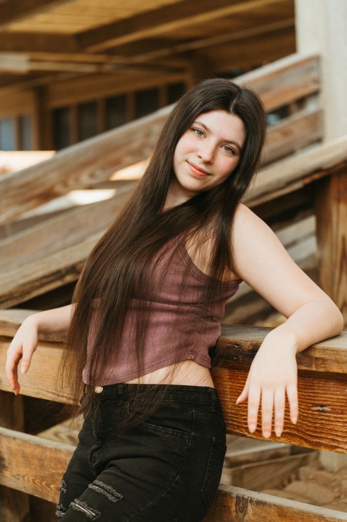 Young woman with long dark hair poses against rustic wooden railings in a mauve crop top and black ripped jeans.
