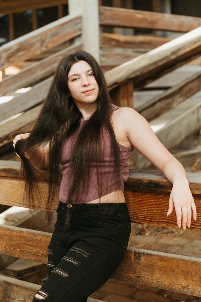 Teen girl with long dark hair sits on rustic wooden steps wearing a sheer mauve crop top and black ripped jeans.