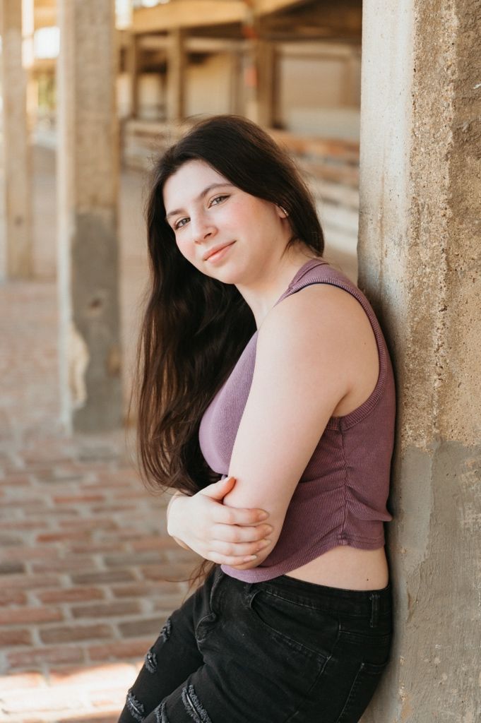 Young woman with long dark hair leans against a stone wall outdoors, wearing a purple crop top and black jeans.