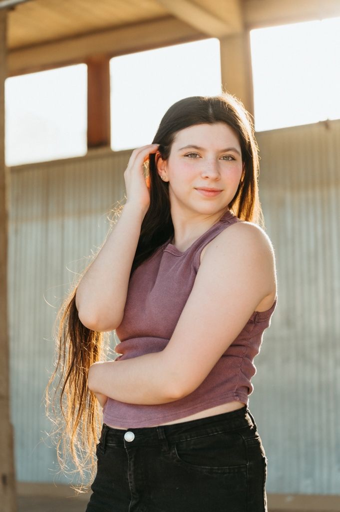 Young woman with long dark hair in purple crop top and black jeans poses outdoors in golden sunlight.
