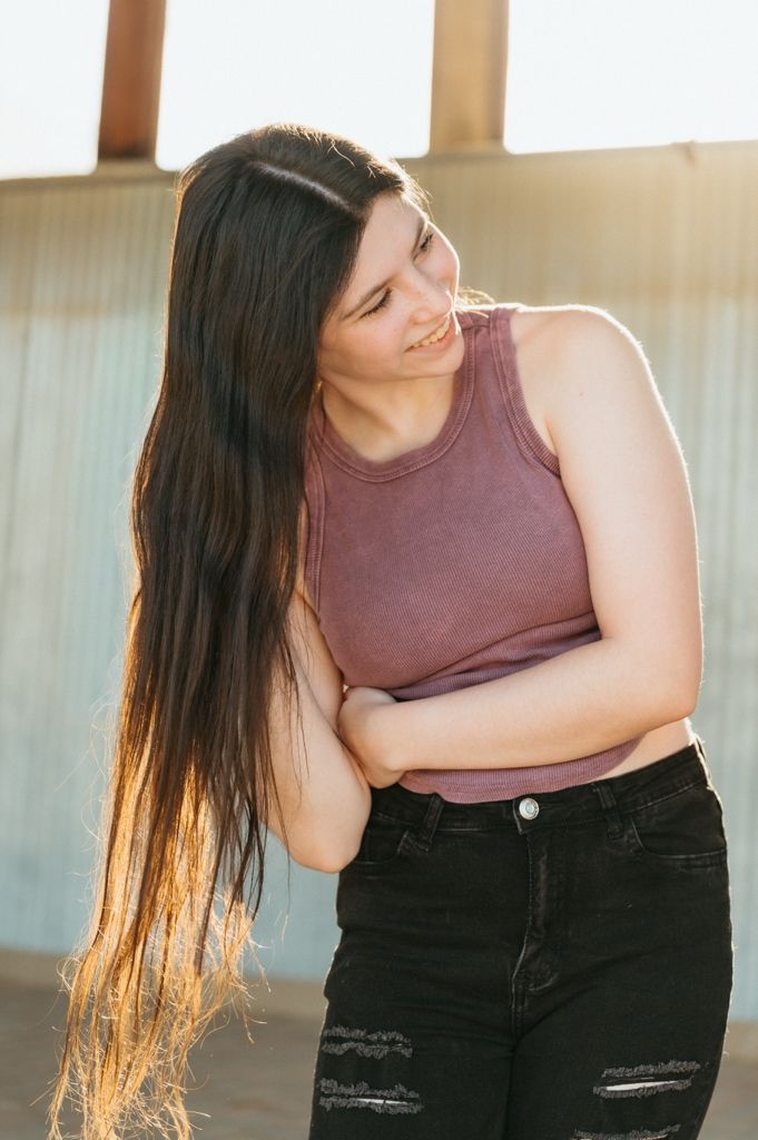 Young woman with long dark hair smiling, wearing a pink tank top and black ripped jeans in golden hour light.