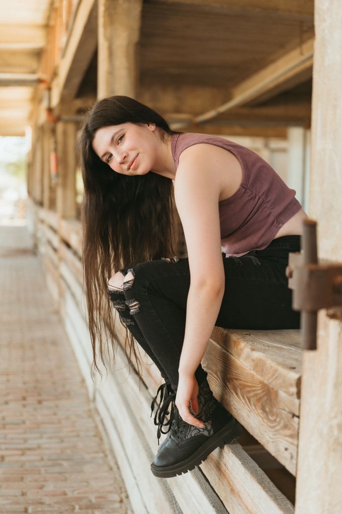Young woman with long dark hair in purple tank top and black jeans adjusting black boots on wooden bleachers.