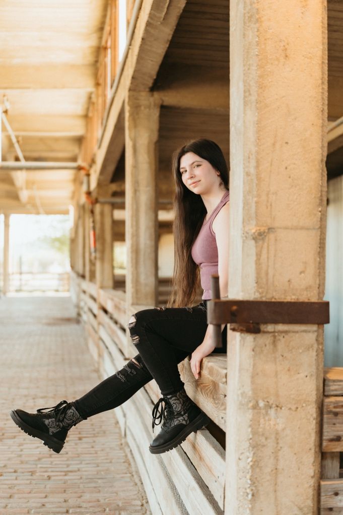 Young woman with long dark hair sits on a ledge wearing a pink top, black jeans, and combat boots in an urban setting.