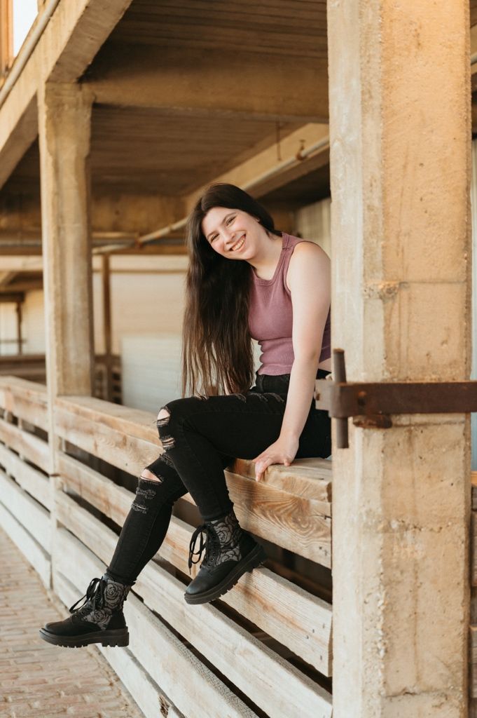 Smiling young woman with long dark hair sitting on wooden bleachers wearing purple tank top and black ripped jeans.