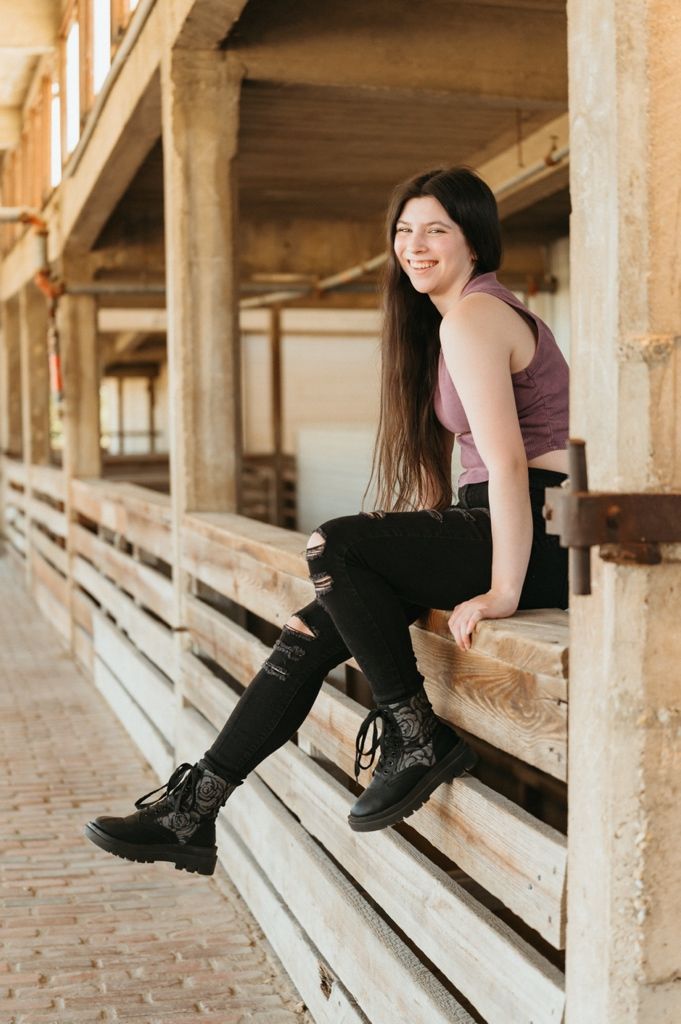 Smiling young woman with long dark hair sits on wooden bleachers wearing ripped black jeans and boots.