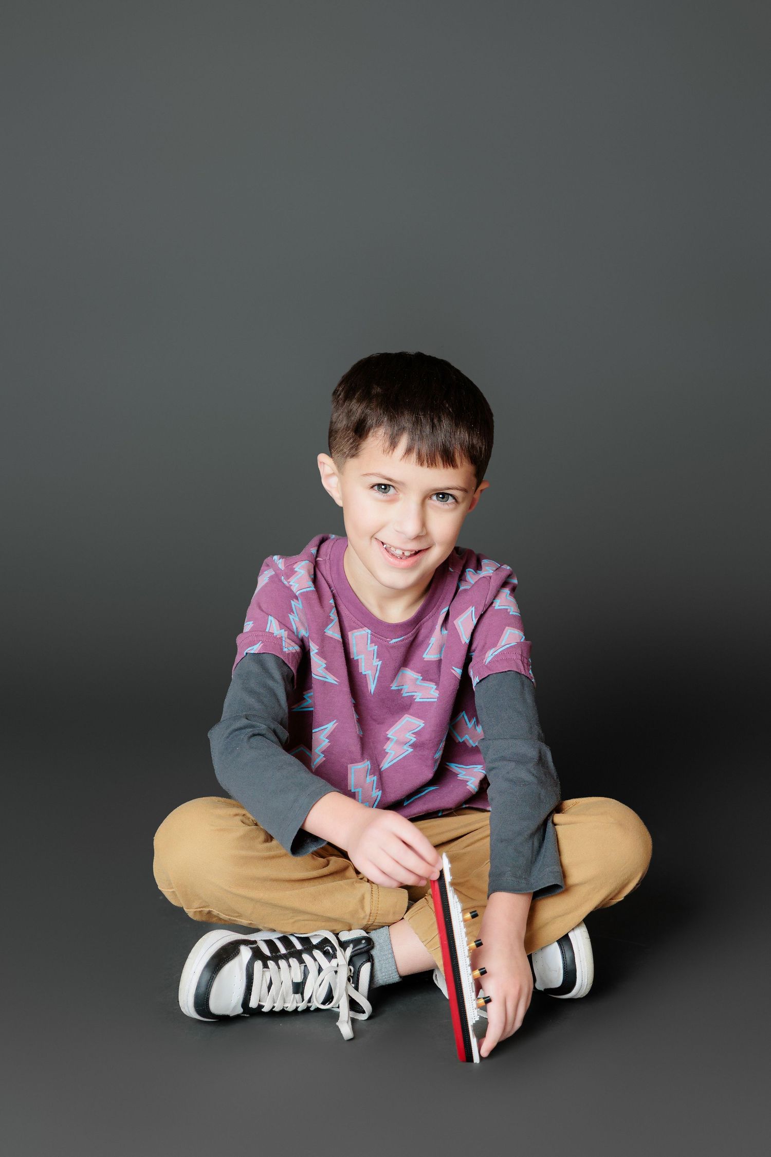 Child in purple sweater and khaki pants sits cross-legged on dark floor with black sneakers.