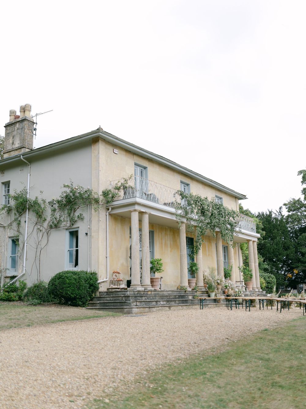 Elegant two-story country manor house with climbing vines, stone columns, and gravel driveway set in lush green grounds.