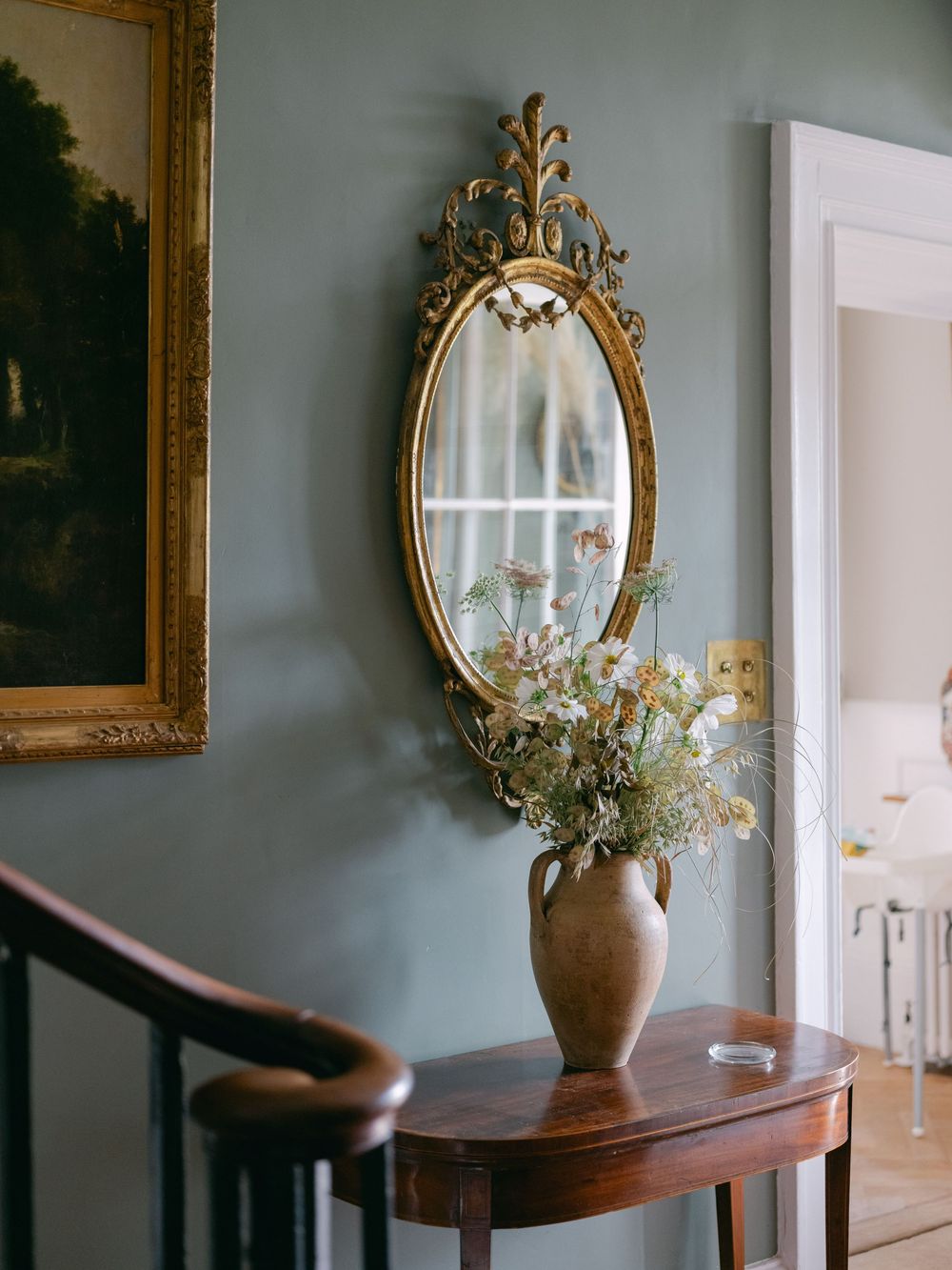 Elegant hallway with ornate gold oval mirror above wooden table with wildflower vase beside staircase on sage green wall.