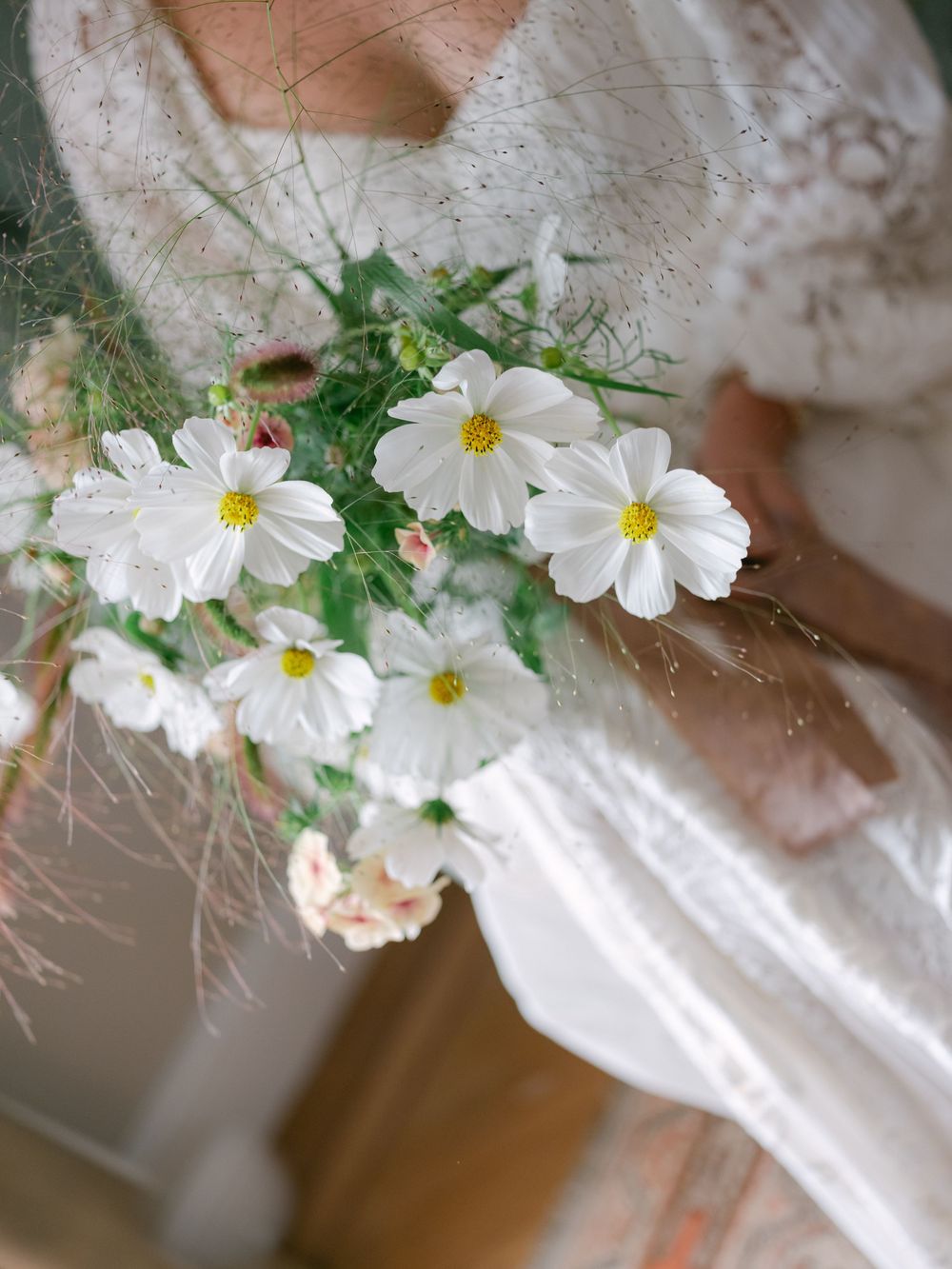 Bride holding a delicate bouquet of white cosmos flowers with yellow centers and wispy green foliage in a lace dress.