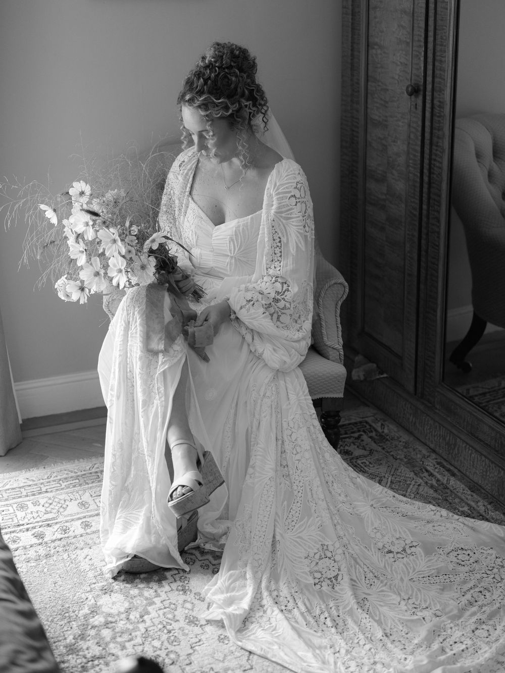 Black and white portrait of curly-haired bride in lace gown seated holding a wildflower bouquet.