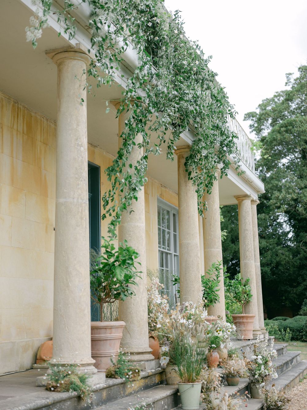 Grand country estate facade featuring tall stone columns draped with cascading green vines and terracotta potted plants.