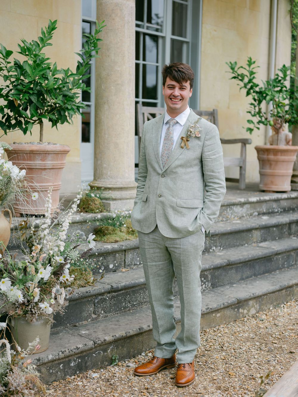 Groom in sage green linen suit with boutonniere smiling on stone steps of elegant venue with potted trees.