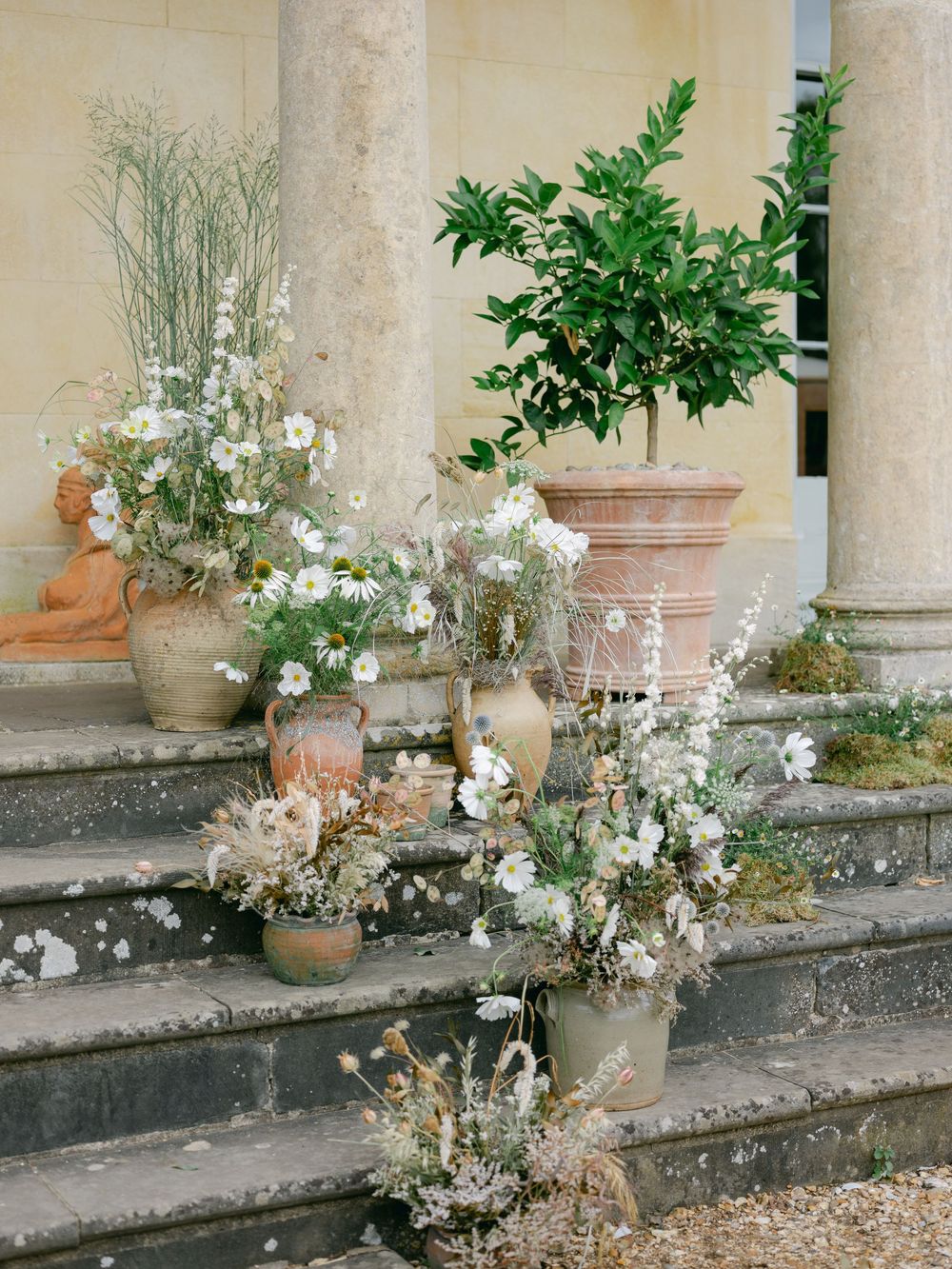 Terracotta pots with white wildflowers and greenery arranged on stone steps of an elegant venue.