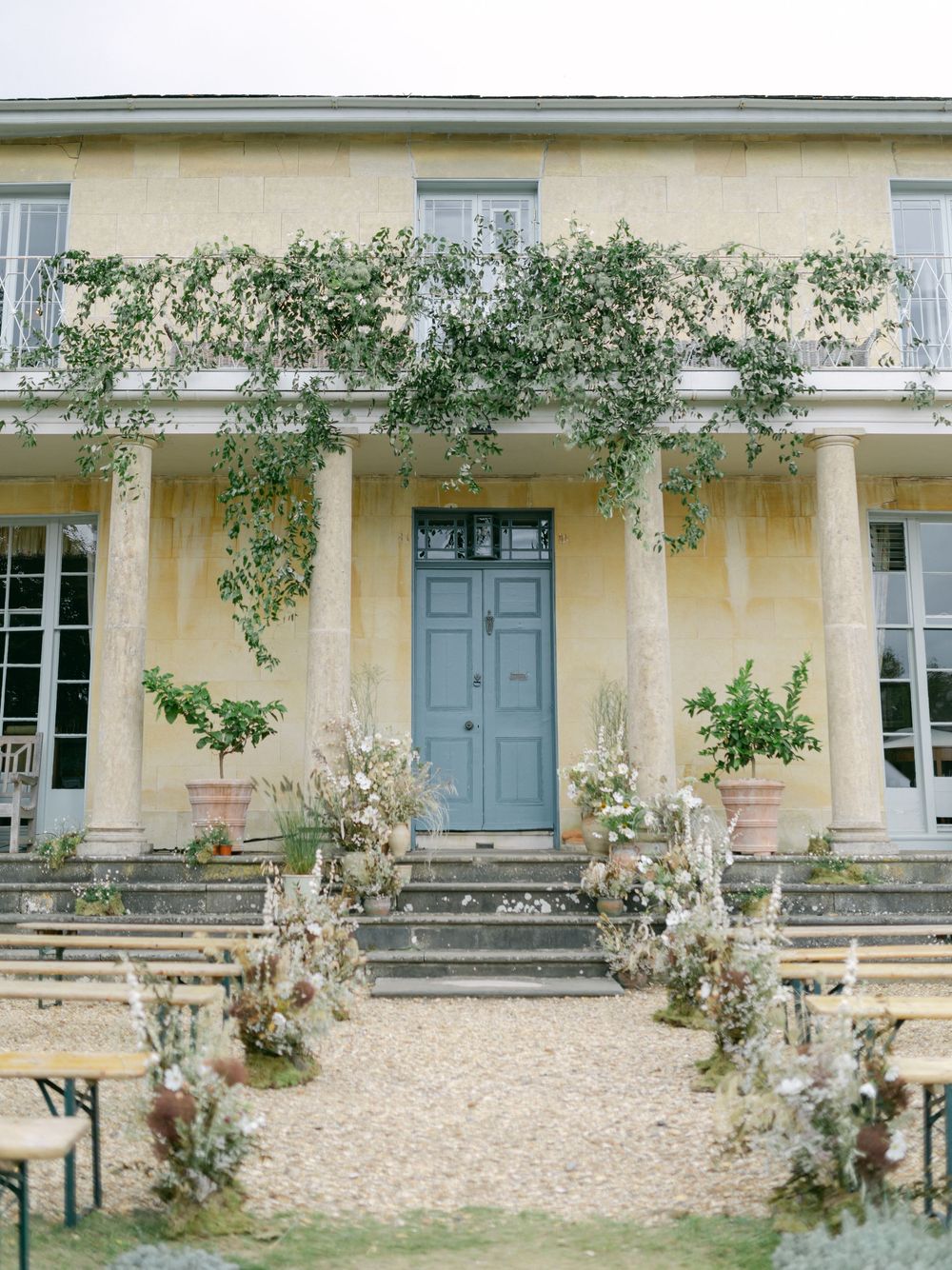 Elegant yellow manor house wedding venue with blue door, climbing vines, floral aisle arrangements, and wooden chairs on gravel.