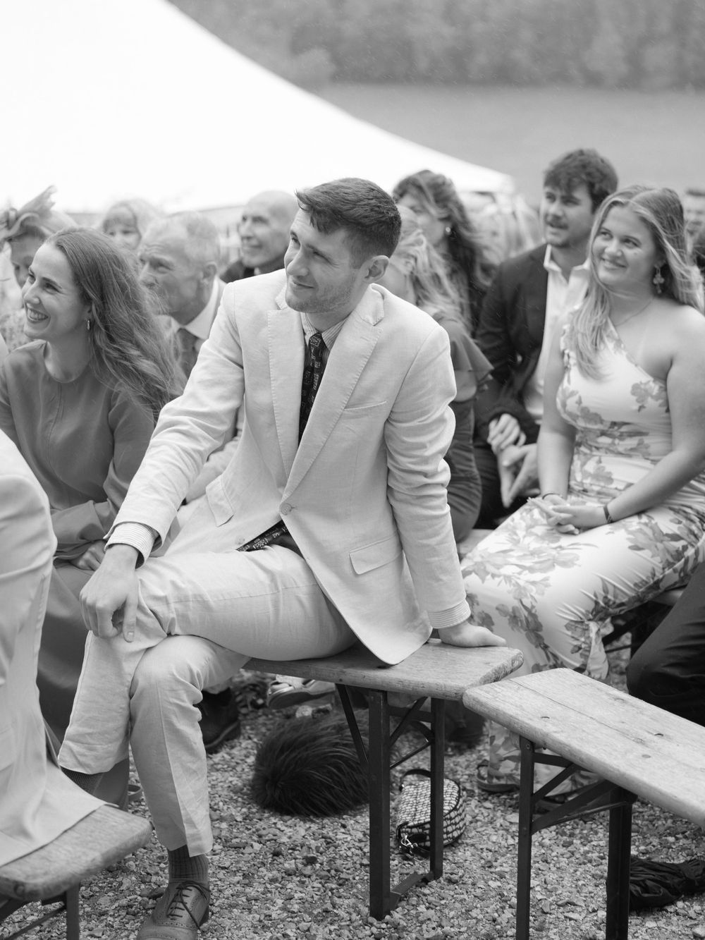 Black and white photo of wedding guests seated on benches under a marquee during an outdoor ceremony.