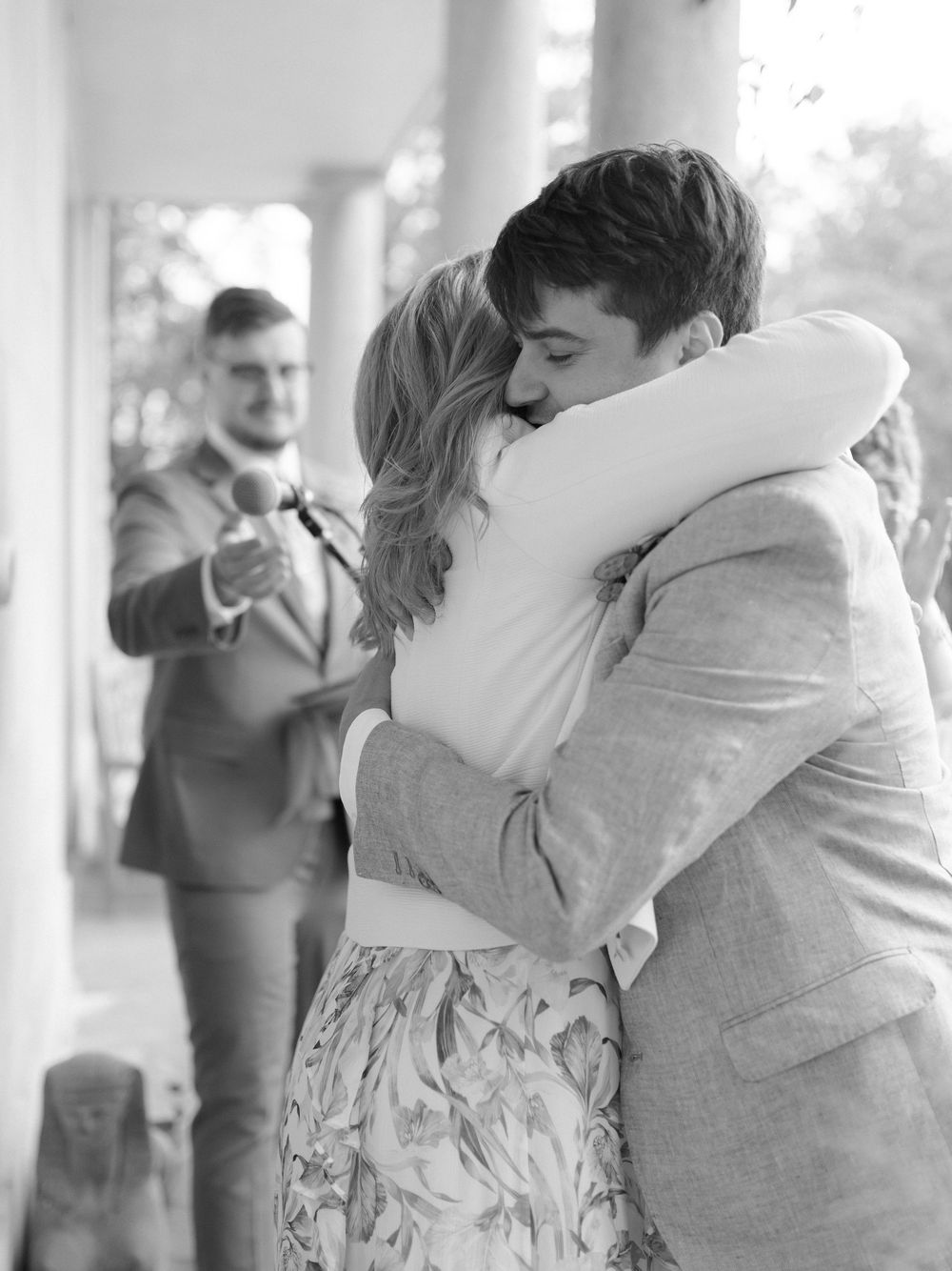 Black and white photo of a couple embracing warmly at a wedding ceremony with a guest applauding in the background.