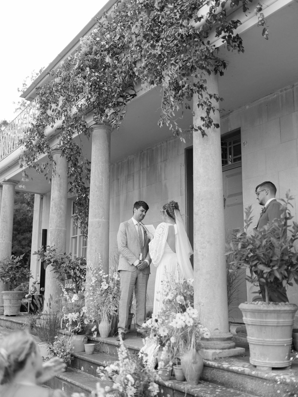 Black and white photo of a couple exchanging vows on steps of a columned estate.