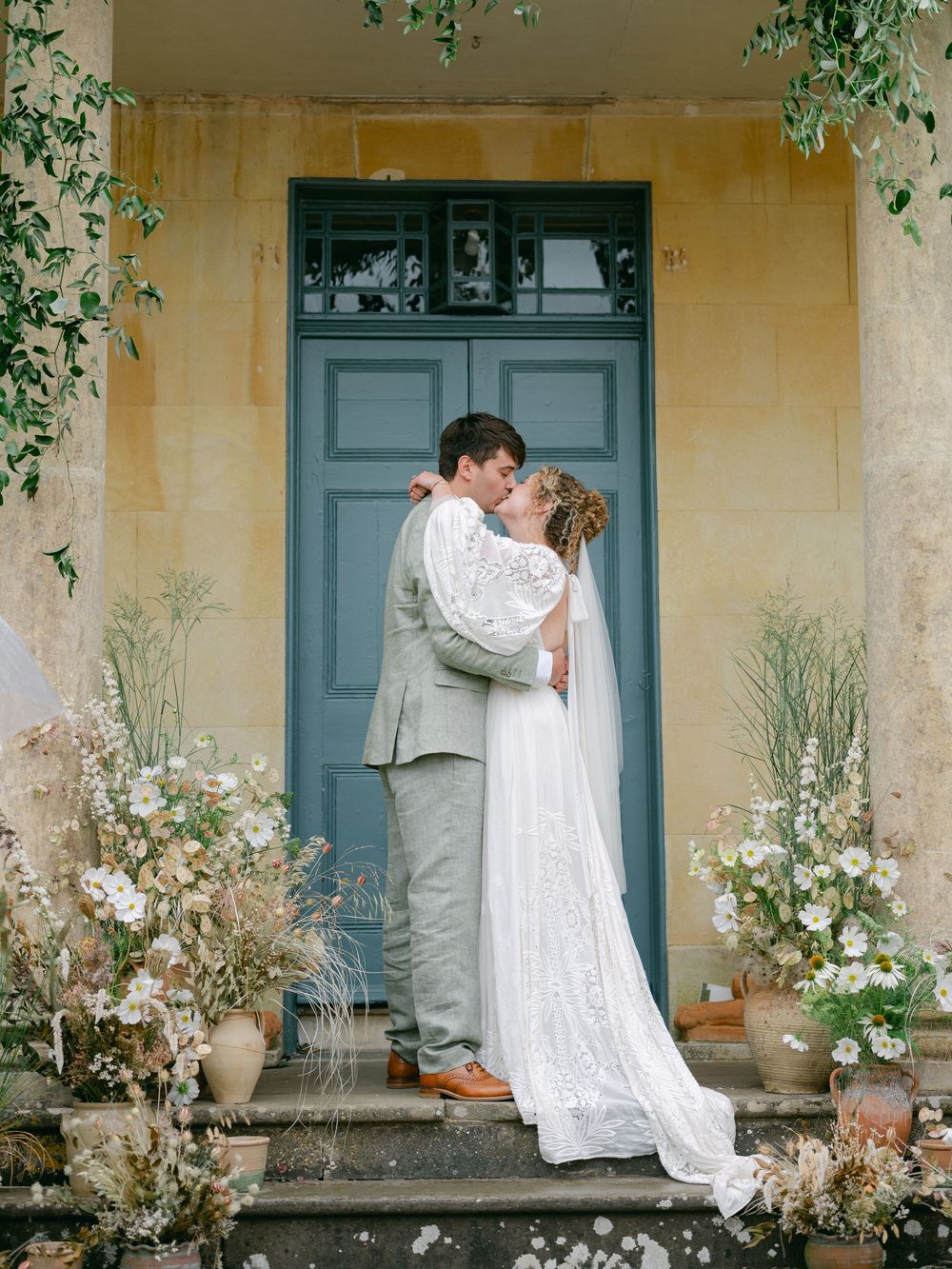 Bride and groom share a kiss on stone steps before a blue door, surrounded by lush floral arrangements.