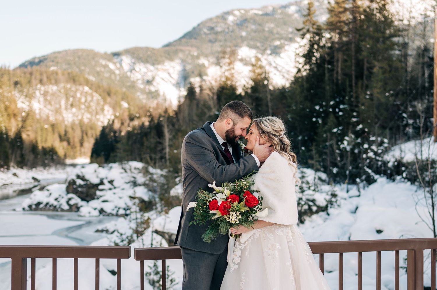 Bride and groom share a romantic winter moment on a snowy mountain deck, bride holding red roses bouquet.