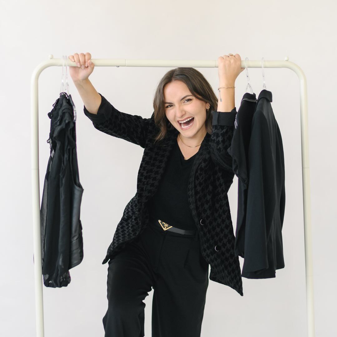 Woman in black outfit joyfully posing with a clothing rack holding dark garments against a white background.