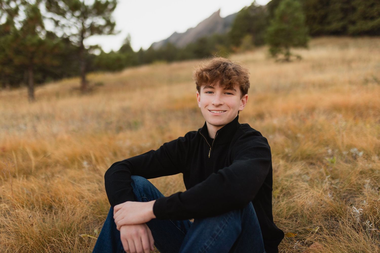 A youth in a black jacket sits in a golden grass field with mountains and pine trees in the background taken by Aurora senior photographer