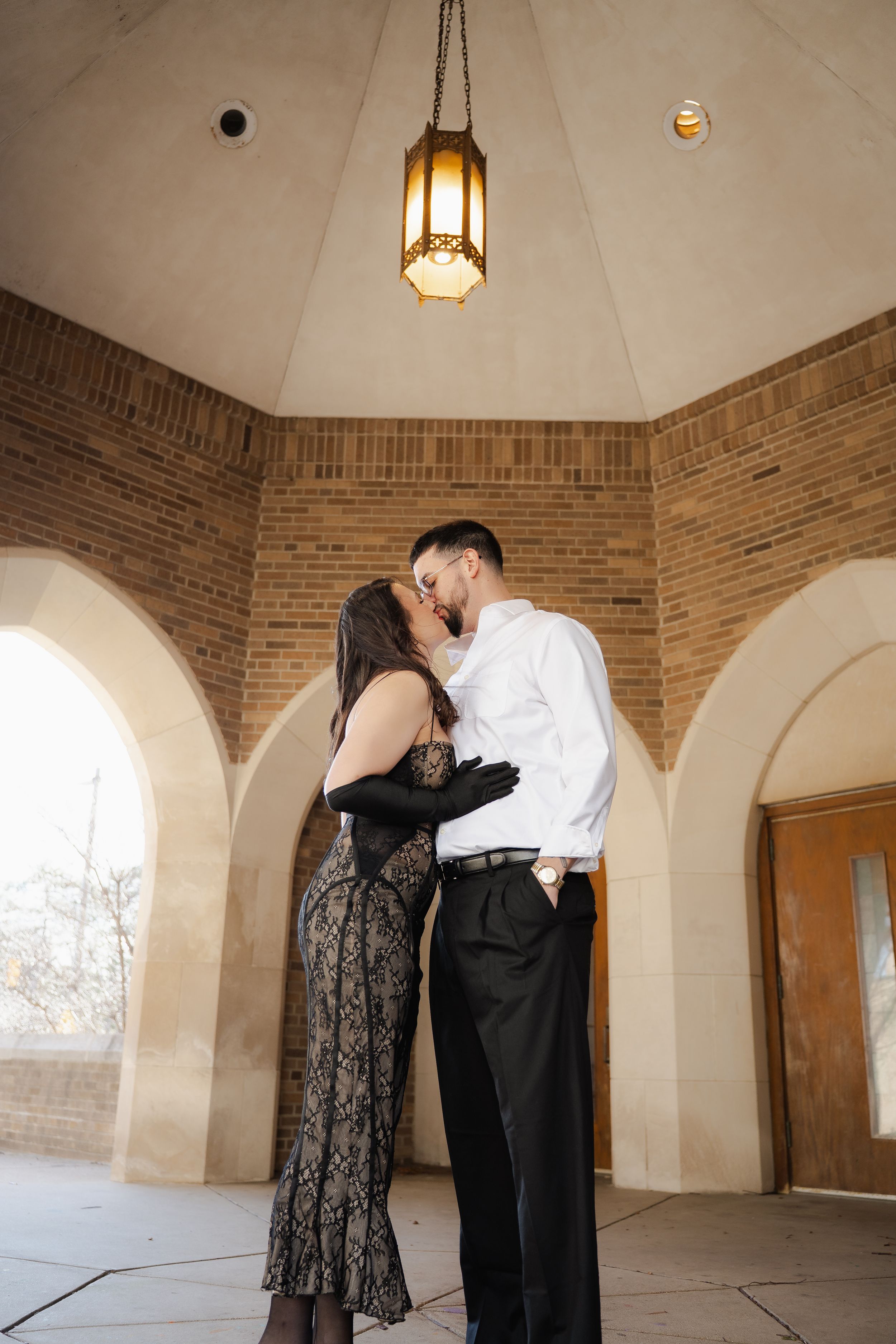 A couple embraces in an elegant brick room with arched windows and pendant lighting creating a romantic atmosphere.