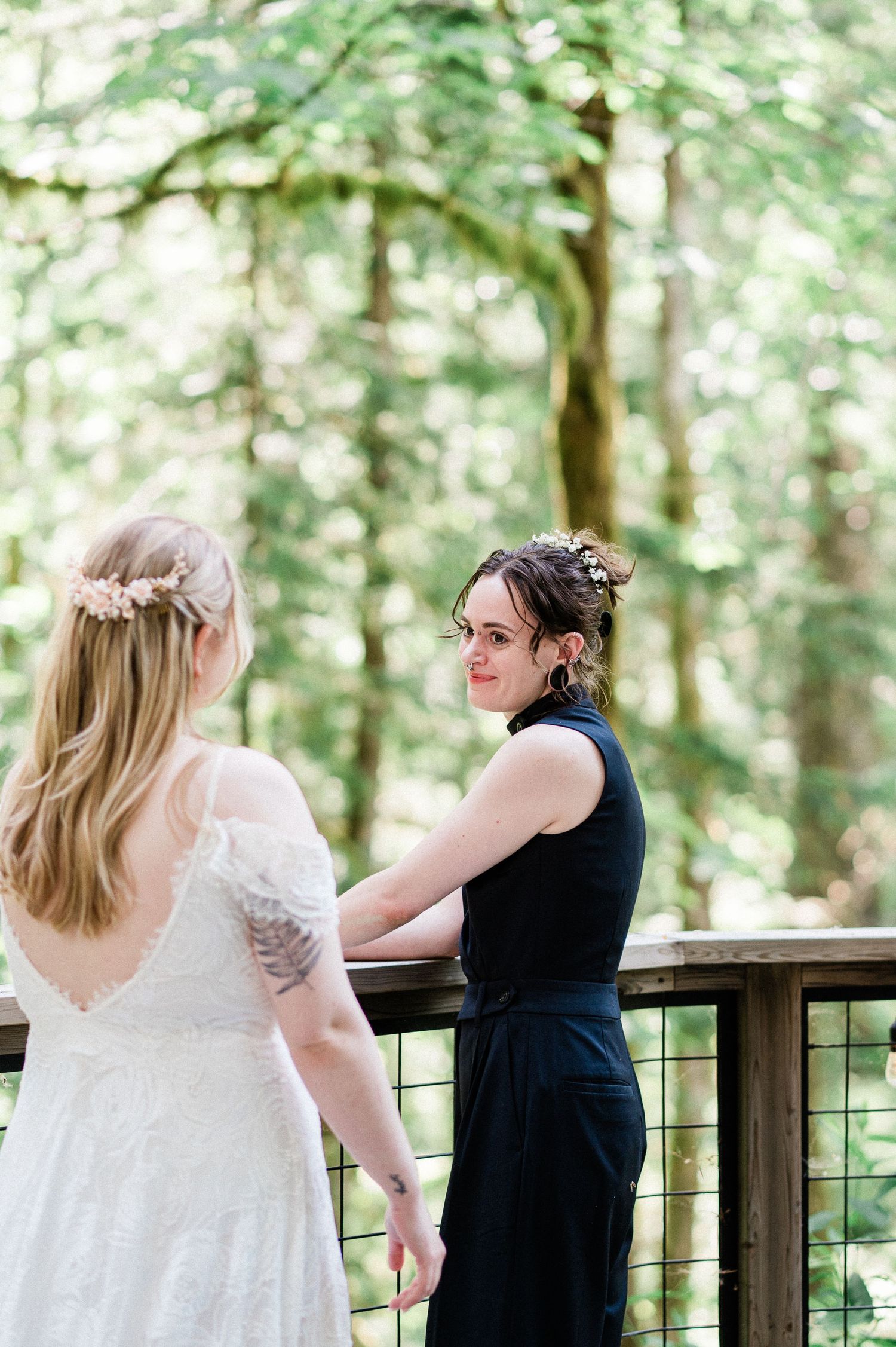 Two brides share a first look moment on a wooden deck surrounded by lush green forest scenery.