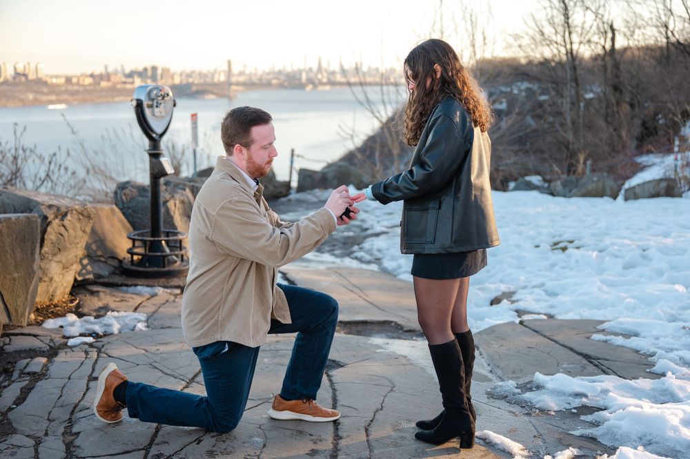 A couple stands on a snowy cliffside during a winter proposal with the city skyline visible in the background in the Palisades in NJ.