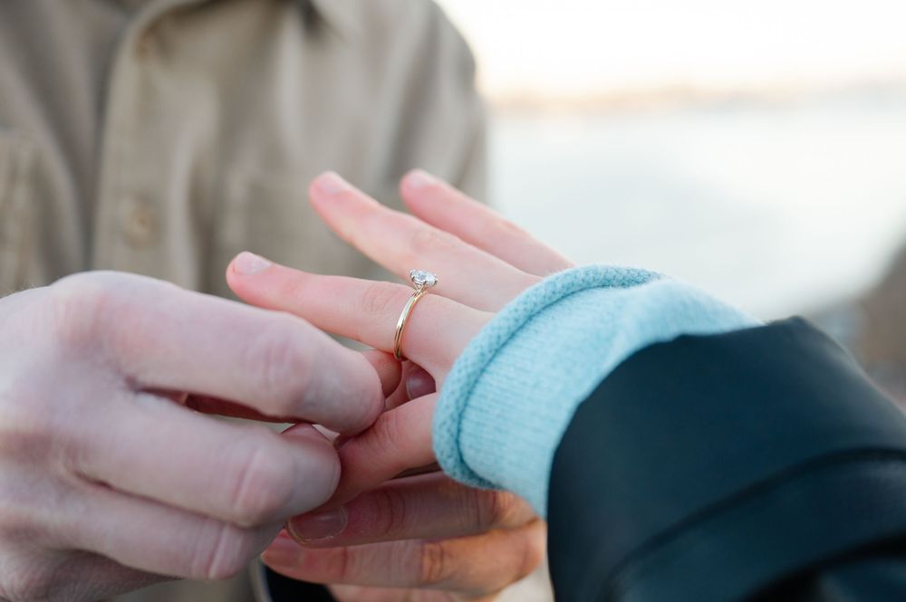 Close-up view of hands exchanging a delicate engagement ring, with one person wearing a light blue sleeve.