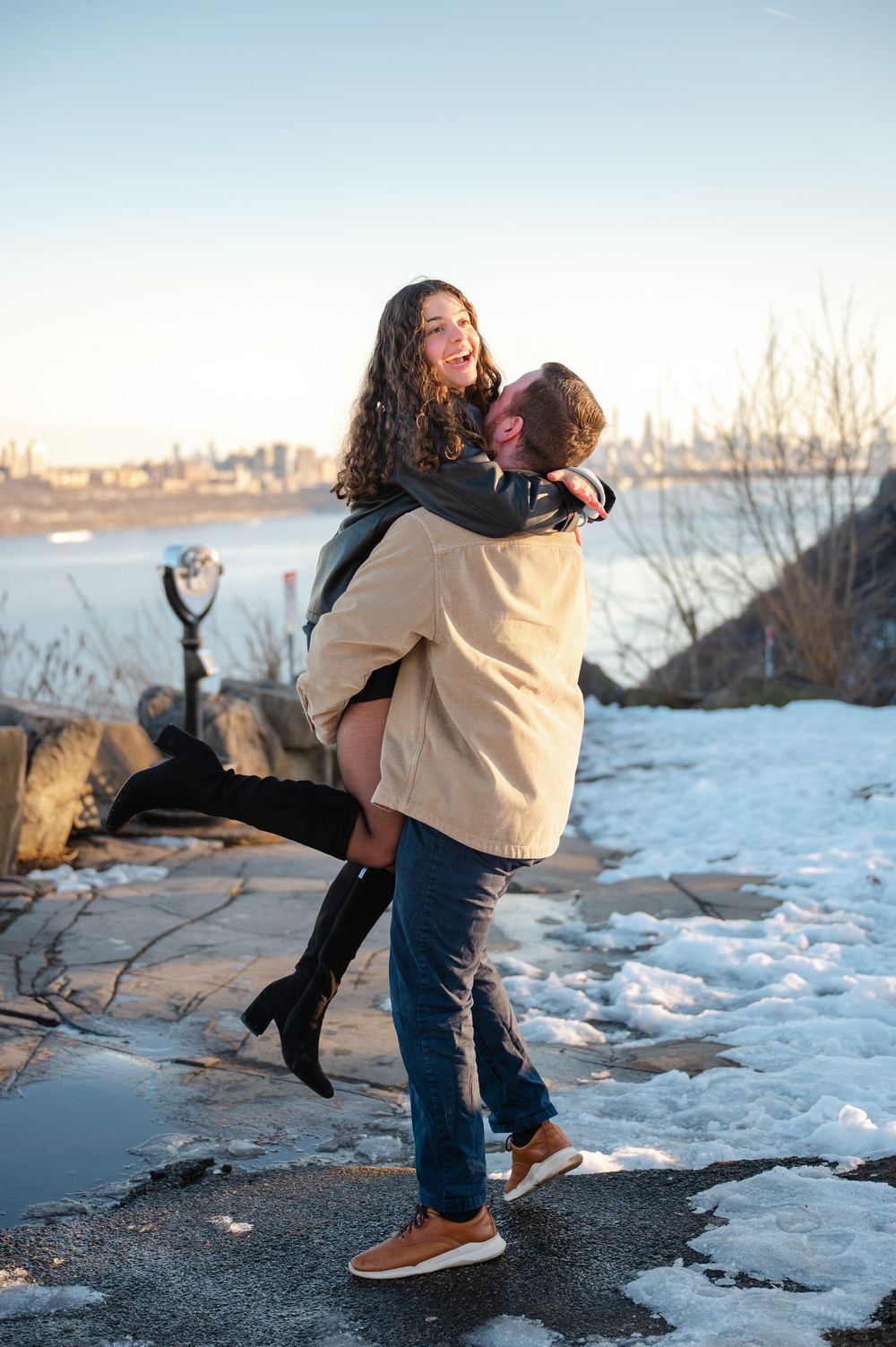 A couple embraces playfully in a winter scene by the water with snow and rocks in the background.