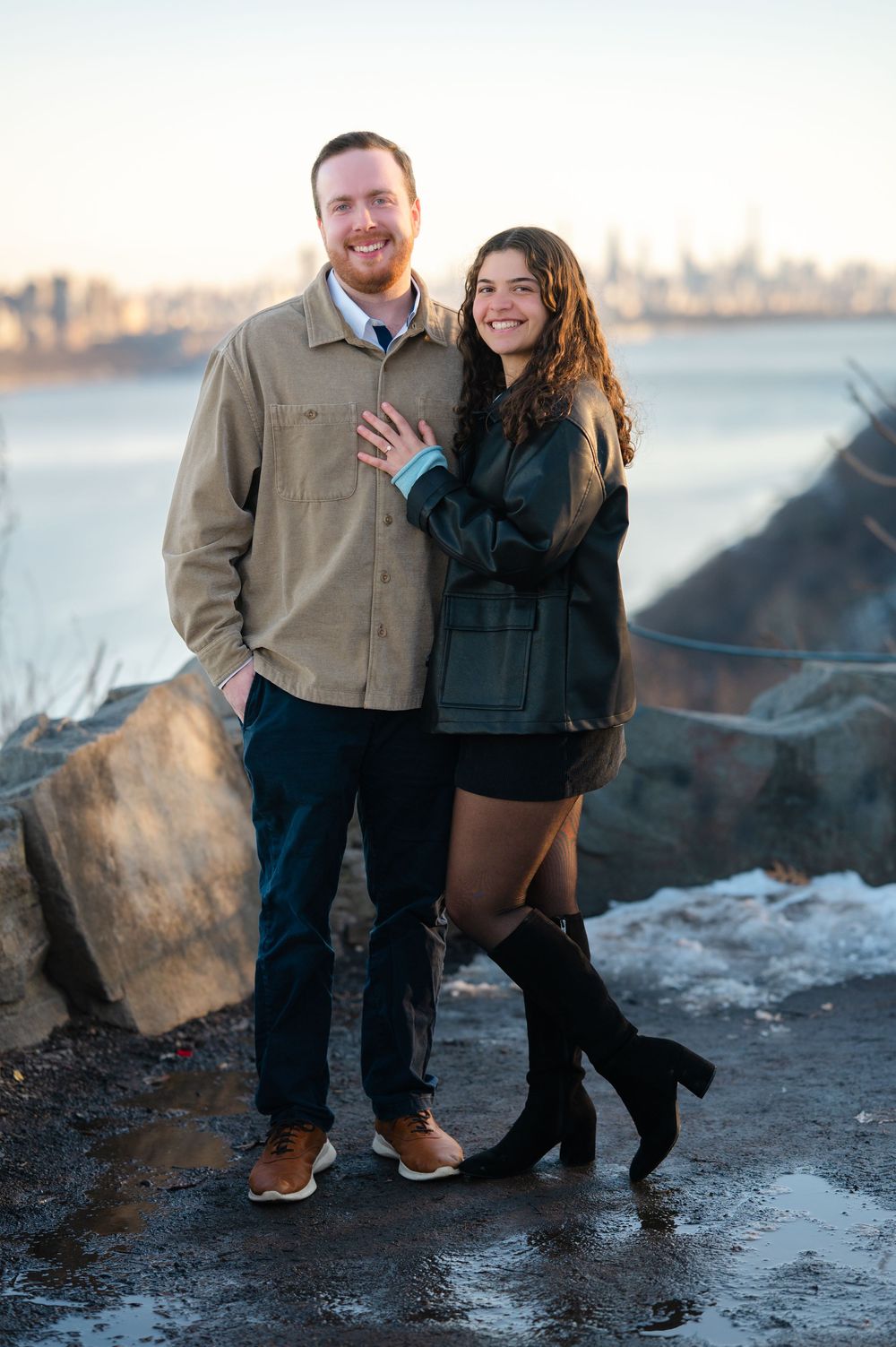 A couple poses together in the evening sunlight with the city skyline in the background during their proposal in the Palisades in New Jersey