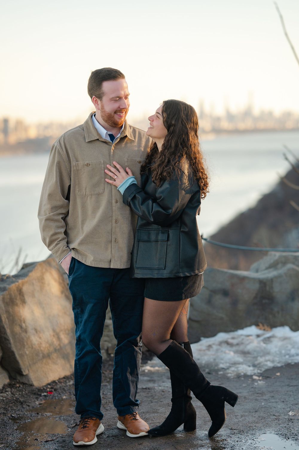 A couple shares a romantic moment along a rocky waterfront during sunset, creating an intimate silhouette against the skyline.