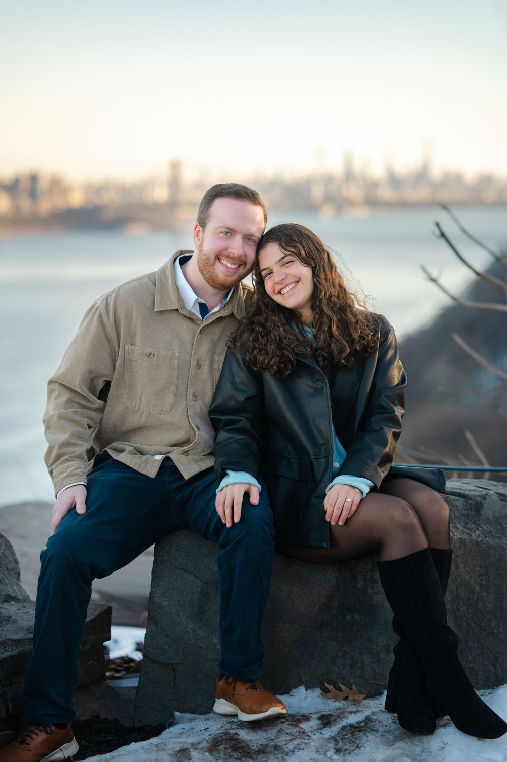 A couple in casual winter attire sits together on rocks by the waterfront during sunset with a city skyline view.