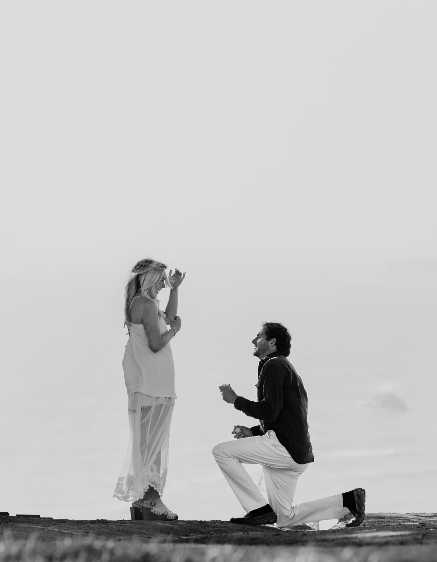 Black and white photo of a surprise proposal moment with one person kneeling on a cliff overlooking the horizon.