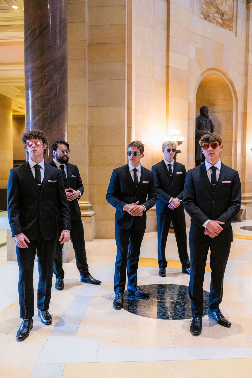 Five men in black suits and sunglasses pose formally in an elegant marble hall.