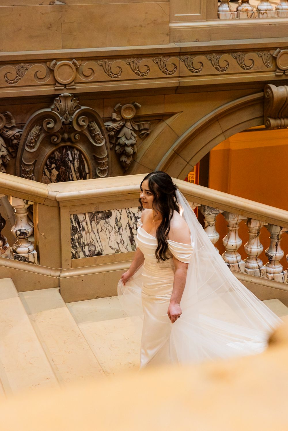 Bride in off-shoulder gown and veil walks up ornate marble staircase in elegant venue.