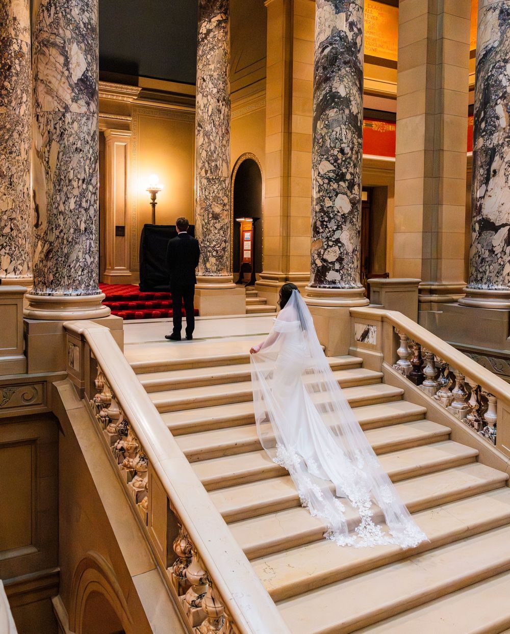 Bride in white gown and veil descends grand marble staircase in elegant venue with ornate columns.